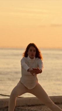 Kadın gün doğumunda denizin yakınındaki beton bir yüzey üzerinde nazikçe esneme egzersizleri yapıyor.