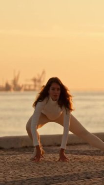 Yoga yapan esnek sporcu kadın şafak vakti asfaltlı bir yüzeyde bir su kütlesinin yanında poz veriyor.