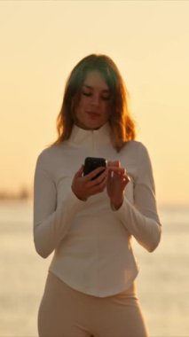 Günbatımında akıllı telefon kullanan kadın ve arka planda şehir manzarası