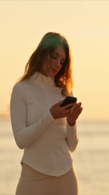 Gün batımında deniz kenarında akıllı telefonunu kullanan genç bir kadın, altın saatin tadını çıkarıyor.