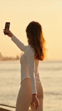 Genç bir kadın gün batımında Tagus nehri ile Lizbon 'da akıllı telefonuyla selfie çekiyor.