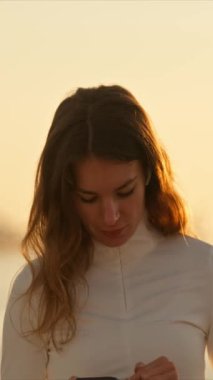 Kulaklık takan ve güneş doğarken deniz kenarında akıllı bir telefon tutan genç bir kadın.