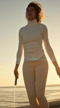 Zinde bir kadın güneş doğarken deniz kenarında atlama ipiyle egzersiz yapıyor.