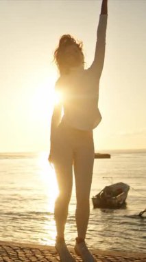 Gün batımında nehrin kenarında dans eden enerjik kadın, altın saatin tadını çıkarıyor.