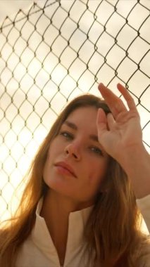 Gün batımında tel örgülerin yanında poz verip saçlarıyla oynayan genç bir kadın.