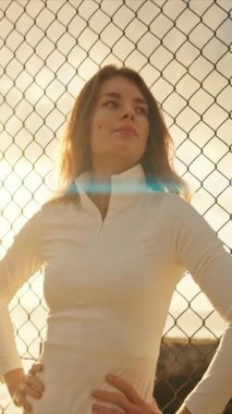 Gün batımında tel örgülerin yanında elleri kalçasında duran genç atletik bir kadın.