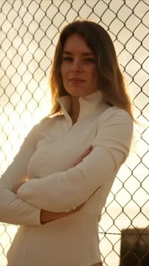 Kadın, arka planda deniz manzaralı ve gün batımlı tel örgülerin yanında dururken kendine güvenir.