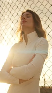 Genç bayan sporcu gün batımında kolları çapraz bir şekilde tel örgüye bağlı duruyor.