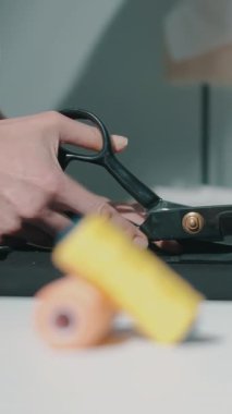 Bir kadın stüdyo ortamında siyah kumaşı makasla keser. İplik makarası ve diğer el işi malzemeleri masada görünür..