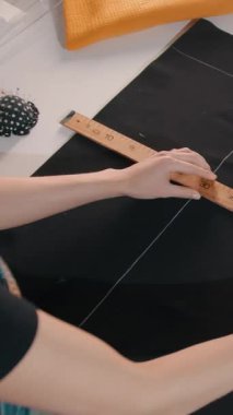 Bir kadın eli cetvelle bir kumaş parçasını ölçüyor. O siyah bir gömlek ve mavi tulum giyiyor.