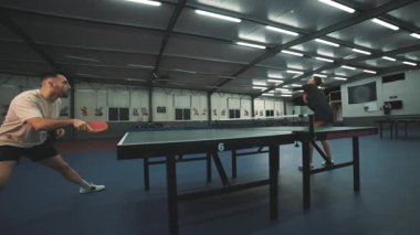 Erkek ve kadın oyuncular spor salonunun içinde dinamik bir masa tenisi oyununda yarışıyorlar.