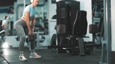 Spor salonunda kulaklıklı, kettlebell bardakları çalan atletik bir kadın.