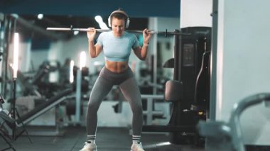 Kulaklıklı atletik bir kadın antrenman sırasında omuzunda halterle çömelme hareketleri yapıyor.