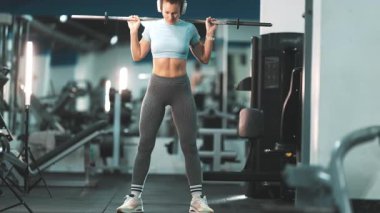 Kulaklıklı kaslı bir kadın halterle egzersiz yapıyor, modern bir fitness kulübünde çömeliyor.