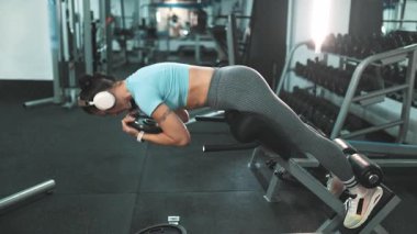 Fit bir kadın, kulaklıklı, belini güçlendiriyor ve kalçasını uzatılmış bir bankta tutuyor.