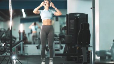 Kulaklık takan atletik bir kadın modern bir spor salonunda halterle ağırlık kaldırıyor.