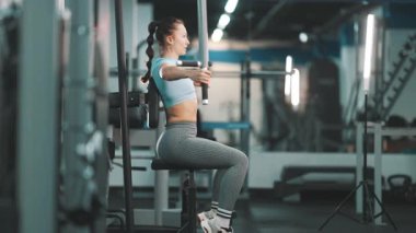 Genç bir kadın fitness kulübündeki kelebek makinesinde göğüs kaslarını çalıştırıyor.