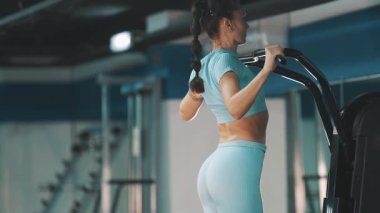 Formda atletik bir kadın egzersiz makinesinde barfiks çekerek sırtını ve kollarını güçlendiriyor.