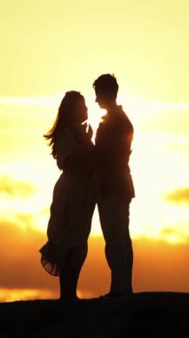 Sevgi dolu bir çiftin silueti bir tepenin üstünde canlı bir günbatımı arkaplanına sarılıyor.