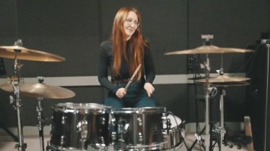 Uzun kızıl saçlı ve gözlüklü genç bir kadın profesyonel bir kayıt stüdyosunda davul çalıyor.
