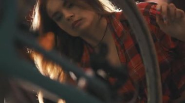 Ekose gömlekli genç bir kadın bir atölyede bisikleti dikkatlice inceliyor ve onarıyor.