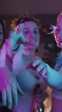 Süslü giysiler içinde güzel yetişkin kadınlar evde parti veriyor. Ellerini kameraya götürüp gülüyorlar. Orta çekim