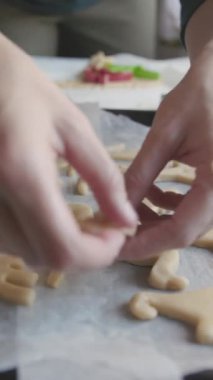 Dişi ellerin, çiğ hamurdan dinozor şekilli kurabiyeleri kesmesi ve kurusıkı mermileri pişirme tepsisine sermesi. Yakın plan.