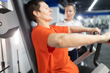 Olgun bir kadın spor salonunda spor yapıyor, kişisel antrenör ona yardımcı oluyor.