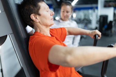 Kıdemli bir kadın arka planda kişisel antrenörüyle spor makinesinde egzersiz yapıyor.