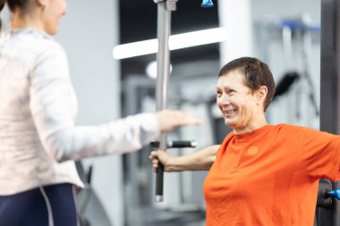 Kıdemli kadın spor yaparken gülümsüyor, bir eğitmenden rehberlik alıyor.