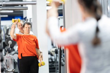 Yaşlı kadın dambılları kaldırıyor, kadın fitness eğitmeninden yardım alıyor.