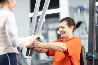 Bir beden eğitimi hocasının rehberliğiyle göğüs presi makinesinde çalışan mutlu bir son sınıf kadını.