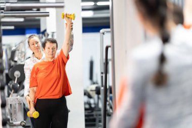 Kıdemli kadın spor salonunda kişisel antrenörünün yardımıyla sarı dambılları kaldırıyor.