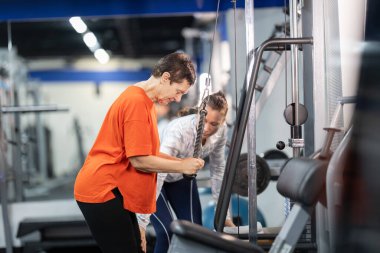 Son sınıf öğrencisi bir kadın, kadın koçundan rehberlik alarak bir kablo makinesinde kol egzersizi yapıyor.