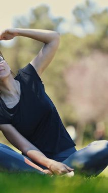 Yetişkin bir kadın doğada esneme hareketleri yapar. Lotus pozisyonunda oturur ve yan tarafa doğru eğilir. Orta çekim