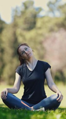 Gözlüklü gülümseyen yetişkin kadın lotus pozisyonunda oturur ve boynunu ısıtır. Orta çekim