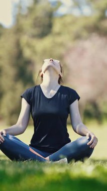 Gözlüklü yetişkin bir kadın parkta lotus pozisyonunda oturur ve boynunu ısıtır. Orta çekim