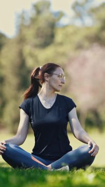 Doğa üzerine yoga yapıyor. Gözlüklü yetişkin bir kadın lotus pozisyonunda oturup boynunu ısıtıyor. Orta çekim