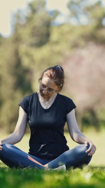 Doğa üzerine yoga yapan gözlüklü bir kadın lotus pozisyonunda oturup boynunu ısıtıyor. Orta çekim