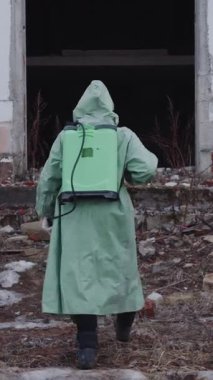 Gaz maskeli ve koruyucu giysili bir adam kimyasal tankın kayışını ayarlayıp terk edilmiş binaya giriyor. Orta çekim