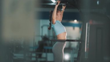 Formda bir kadın bir makinede destekli barfiks çekiyor, sırt ve kol kaslarını güçlendiriyor.