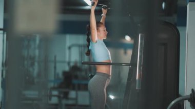 Formda genç bir kadın spor salonunda destekli barfiks çekiyor.