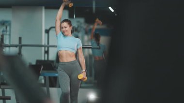 Modern bir fitness kulübünde dambıllarla omuz egzersizi yapan sağlıklı bir kadın.