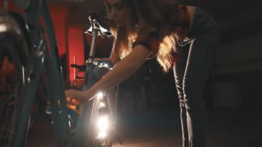 Ekose gömlekli genç bir kadın loş ışıklı, atmosferik garajda bisikleti dikkatlice siliyor.