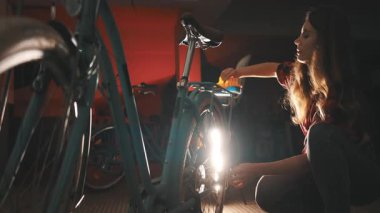 Adanmış kadın bisikletçi loş bir garajda gece gezintisi için klasik mavi bisikletini hazırlıyor.