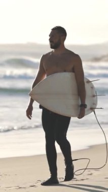 Çıplak gövdeli bir sörfçü deniz kıyısında sörf tahtasıyla yürüyor. Orta vuruş.