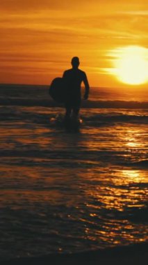 Gün batımında deniz dalgaları arasında giden atletik bir sörfçünün silueti. Orta çekim