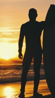 Gün batımında sörf tahtasıyla deniz kıyısında duran bir adamın silueti. Orta çekim