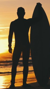 Gün batımında sörf tahtasıyla deniz kıyısında duran atletik bir adamın silueti. Orta çekim