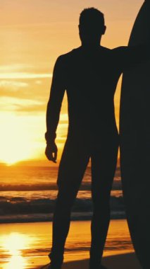 Bir sörfçünün silueti elinde sörf tahtasıyla deniz kenarında gün batımını seyrediyor. Orta vuruş.